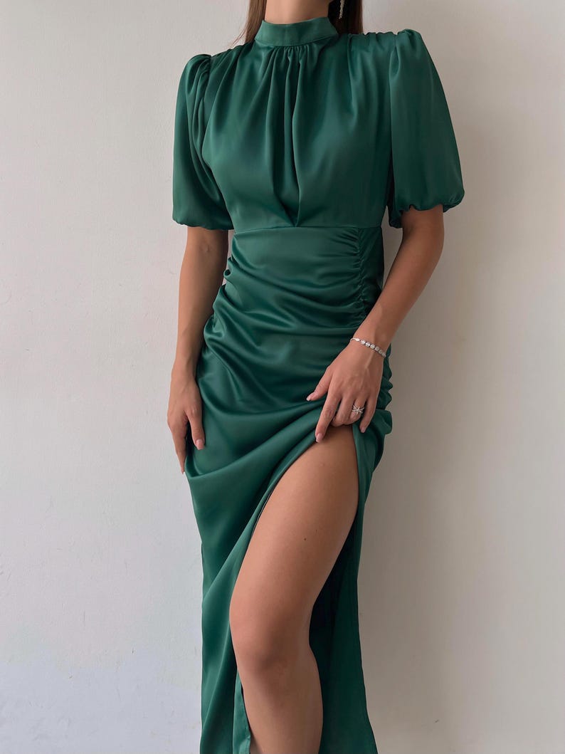 May include: Emerald green satin dress with a high neckline, short puff sleeves, and a thigh-high slit. The dress features ruching on the side and a flowing skirt. A sophisticated formal wear option.