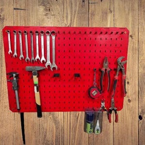 Può includere: Una pannello forato rosso con vari attrezzi manuali, tra cui chiavi inglesi, pinze, un martello e un metro a nastro. Gli attrezzi sono ben organizzati, suggerendo un'officina. Il pannello è montato su una superficie di legno.