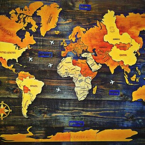 May include: Wooden world map with countries in shades of yellow and orange, mounted on a dark blue and brown wooden background. Includes small airplane cutouts and country labels. A compass rose is in the lower left corner.