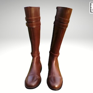 Puede incluir: Un par de botas altas de cuero marrón. Cada bota presenta una puntera redondeada, detalles de correas decorativas alrededor de la pantorrilla y cierre de cremallera. Las botas se muestran sobre un fondo blanco.