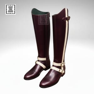 Puede incluir: Un par de botas de montar altas, de color burdeos, con detalles en crema. Las botas tienen una franja blanca en el lateral y correas color crema con hebillas doradas. Las botas tienen una cremallera y el texto "Craft Edge Goods".
