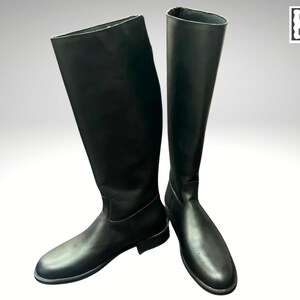 May include: A pair of tall, black leather riding boots. The boots are knee-high with a smooth, polished finish. The boots are standing upright, showcasing their sleek design and construction. The Craft Edge Goods logo is in the upper right corner.