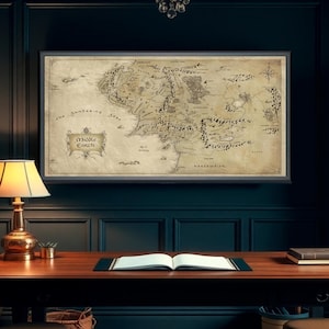Pode incluir: Um mapa emoldurado da Terra-média está pendurado acima de uma mesa de madeira em um escritório. O mapa é bege com texto e ilustrações pretas. Uma luminária de mesa, livros e um banquinho estão em frente à mesa. A sala tem paredes azul-escuras e um lustre.