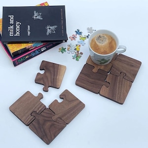 Könnte beinhalten: Vier Untersetzer aus Holz in Puzzleform in dunkelbrauner Farbe. Die Untersetzer sind quadratisch angeordnet, mit einer Tasse Kaffee darauf. Die Untersetzer sind aus Walnussholz gefertigt und haben eine natürliche Holzoptik.