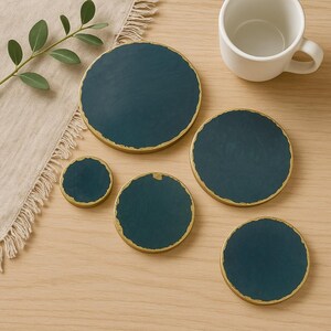 May include: Set of five round coasters with a dark teal blue centre and a gold rim. The coasters are arranged on a light wood surface.