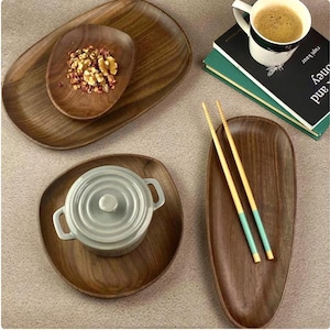 Könnte beinhalten: Eine Sammlung von Holzserviertabletts und -schalen. Ein Tablett enthält einen kleinen grauen Keramiktopf mit Deckel. Ein weiteres Tablett hat eine kleinere Schüssel mit Nüssen und Gewürzen. Eine Tasse Kaffee steht auf einem Stapel Bücher.