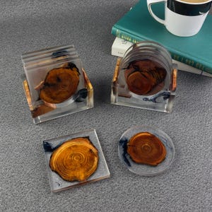 May include: A set of four resin coasters featuring wood slice inserts. The coasters are clear with a wood grain design and a touch of blue. Two are square, two are round. The coasters are displayed on a grey surface.