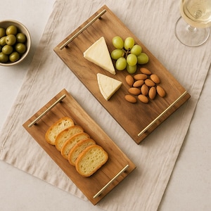 Könnte beinhalten: Zwei rechteckige Serviertabletts aus Holz mit goldenen Griffen. Eines enthält geschnittenes Brot, das andere Käse, Trauben und Mandeln. Eine Schale mit Oliven und ein Glas Weißwein deuten auf einen Snack oder eine Vorspeise hin.