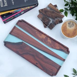 May include: A rectangular wooden serving tray with a light blue resin inlay. The tray has a dark wood grain pattern and is accompanied by matching coasters. A cup of coffee and books are also in the image.