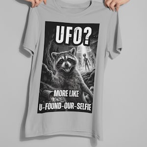 May include: A light grey t-shirt with a black and white graphic. The graphic shows a raccoon with aliens and the text "UFO? MORE LIKE U-FOUND-OUR-SELFIE". The raccoon is smiling in a forest setting.