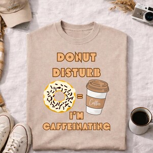 Puede incluir: Camiseta beige con el texto marrón "DONUT DISTURB = I'M CAFFEINATING". El diseño incluye una rosquilla y una ilustración de taza de café. También se ven una gorra de béisbol, una cámara y zapatos.