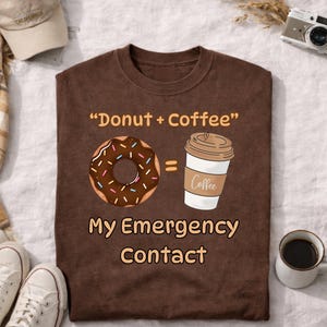 Könnte beinhalten: Ein braunes T-Shirt mit dem Text "Donut + Coffee" über einer Abbildung eines Donuts und einer Kaffeetasse. Darunter steht der Text "My Emergency Contact". Eine Baseballkappe und Turnschuhe sind im Hintergrund.