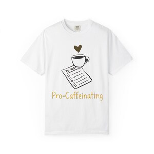 Puede incluir: Camiseta blanca con un gráfico negro y marrón de una taza de café, un corazón y una lista de tareas. El texto "Pro-Caffeinating" está impreso en dorado. La camiseta es de material suave.