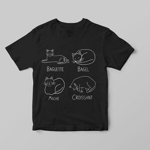 May include: Black t-shirt with white line drawings of cats in various poses, each labeled with a type of bread: Baguette, Bagel, Miche, and Croissant. The shirt is displayed on a light gray background.