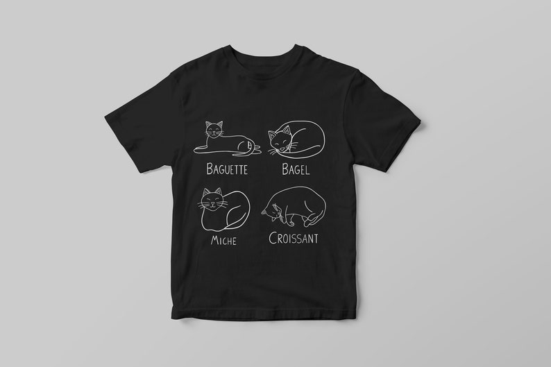 May include: Black t-shirt with white line drawings of cats in various poses, each labeled with a type of bread: Baguette, Bagel, Miche, and Croissant. The shirt is displayed on a light gray background.
