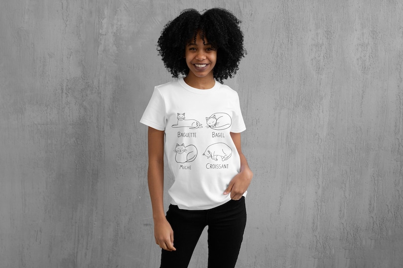 May include: White t-shirt featuring four cat illustrations, each labeled with a different type of bread: Baguette, Bagel, Miche, and Croissant. The shirt is worn by a person with dark, curly hair and black pants.