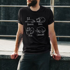 May include: Black t-shirt with a white graphic of four cats in different poses, each labeled with a type of bread: Baguette, Bagel, Miche, and Croissant. The shirt is made of a soft material and is suitable for casual wear.