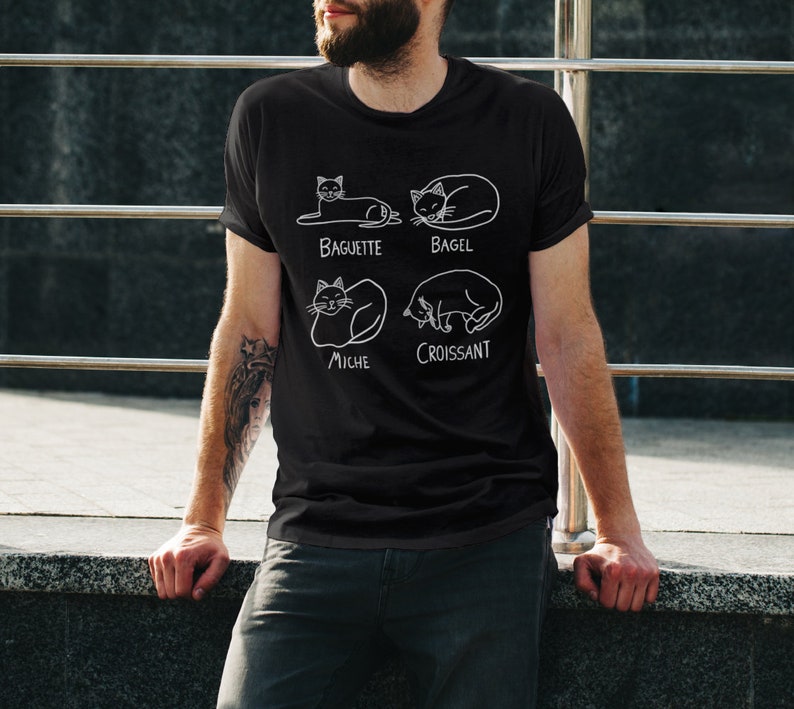 May include: Black t-shirt with a white graphic of four cats in different poses, each labeled with a type of bread: Baguette, Bagel, Miche, and Croissant. The shirt is made of a soft material and is suitable for casual wear.