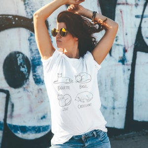 May include: White t-shirt with a black line drawing of four cats, each labeled with a type of bread: Baguette, Bagel, Loaf, and Croissant. The shirt is worn by a person wearing sunglasses.