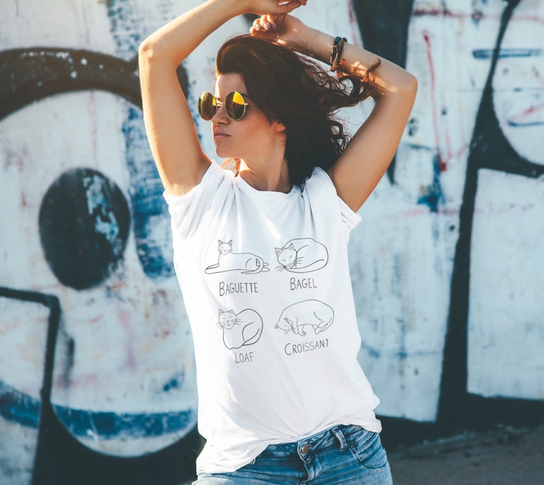 May include: White t-shirt with a black line drawing of four cats, each labeled with a type of bread: Baguette, Bagel, Loaf, and Croissant. The shirt is worn by a person wearing sunglasses.