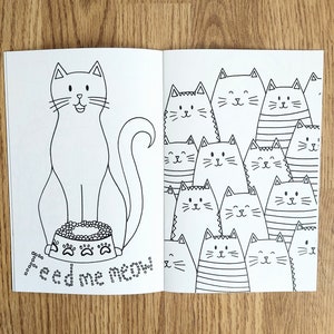 May include: A black and white coloring book featuring a cat with a bowl of food on the left page and a group of cats on the right page.