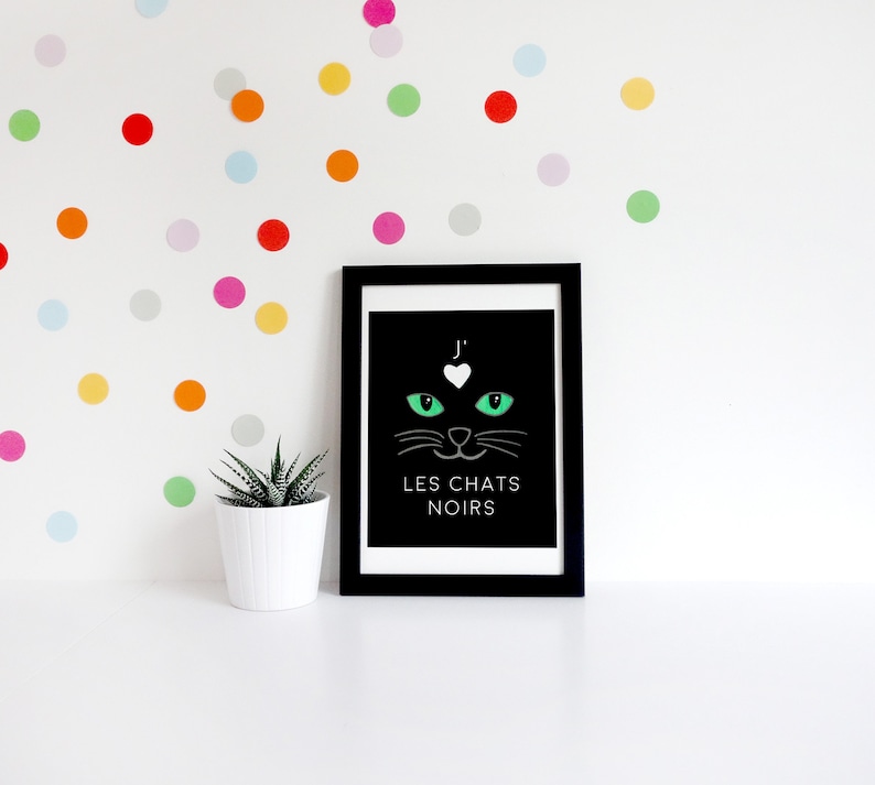 May include: Black and white print featuring a green cat face and the text "J' LES CHATS NOIRS" in a black frame. The print is displayed on a white wall with colourful polka dots.