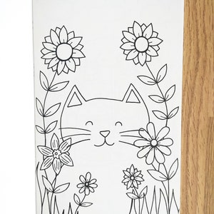 May include: A black and white line drawing of a cat's face surrounded by flowers. The cat has a happy expression with closed eyes and a smile. The flowers are simple designs with petals and stems.