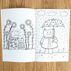 May include: A black and white coloring book page with two cats. One cat is holding an umbrella with stars on it and is standing in the rain. The other cat is standing in a field of flowers.