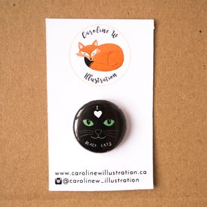 May include: Black and green button with a black cat face and the text "I love black cats". The button is on a white card with the text "Caroline W Illustration" and a website address.