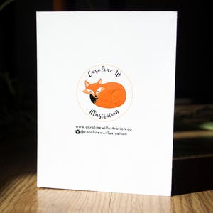 May include: A white card with a logo featuring a sleeping orange fox with a black tail inside a yellow circle. The text "Caroline W Illustration" is above the fox and the text "www.carolinewillustration.ca @carolinew_illustration" is below the fox.