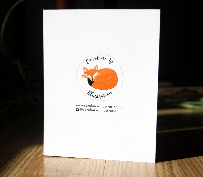 May include: A white card with a logo featuring a sleeping orange fox with a black tail inside a yellow circle. The text "Caroline W Illustration" is above the fox and the text "www.carolinewillustration.ca @carolinew_illustration" is below the fox.