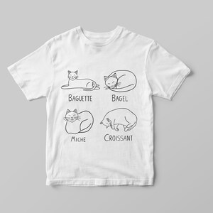 May include: White t-shirt featuring four line-drawn cats in various poses, each labeled with a type of bread: Baguette, Bagel, Miche, and Croissant. The design is in black ink. The shirt is displayed on a light gray background.