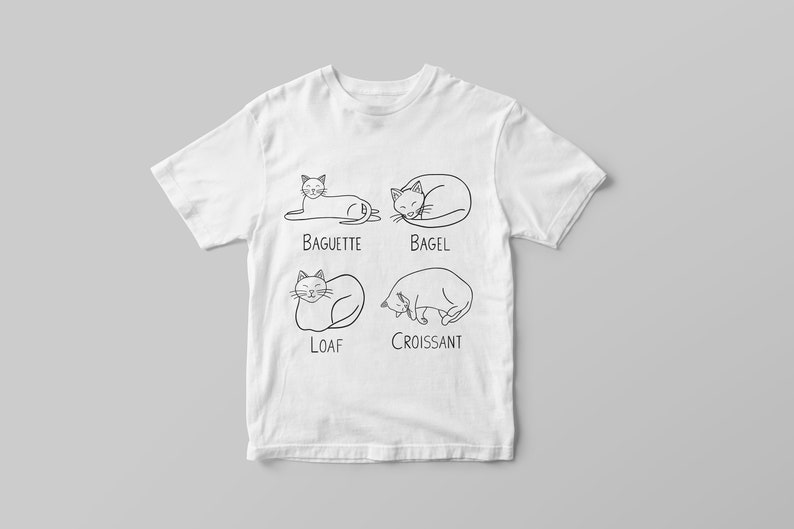 May include: White t-shirt featuring four cat illustrations, each labeled with a type of bread: Baguette, Bagel, Loaf, and Croissant. The black line art depicts cats in various sleeping positions. The shirt is displayed on a gray background.