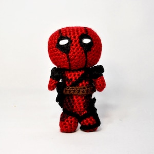 May include: A crocheted Deadpool doll with a red and black suit, a brown belt, and white eyes.