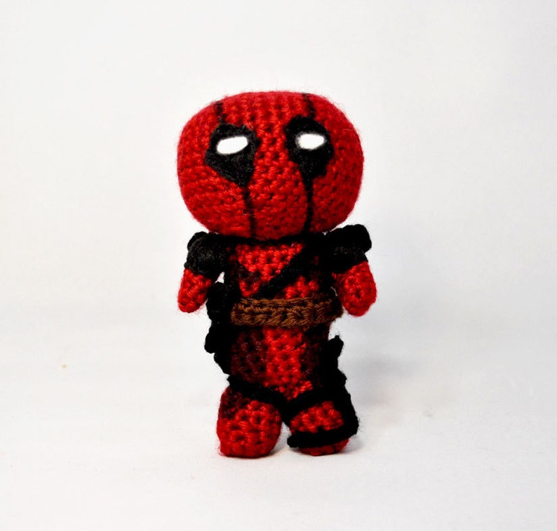 May include: A crocheted Deadpool doll with a red and black suit, a brown belt, and white eyes.
