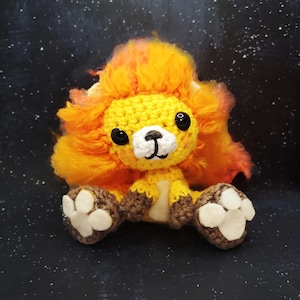 May include: A crocheted lion plush toy with a bright orange mane, yellow body, and white face. The toy has brown crocheted paws with white paw pads.
