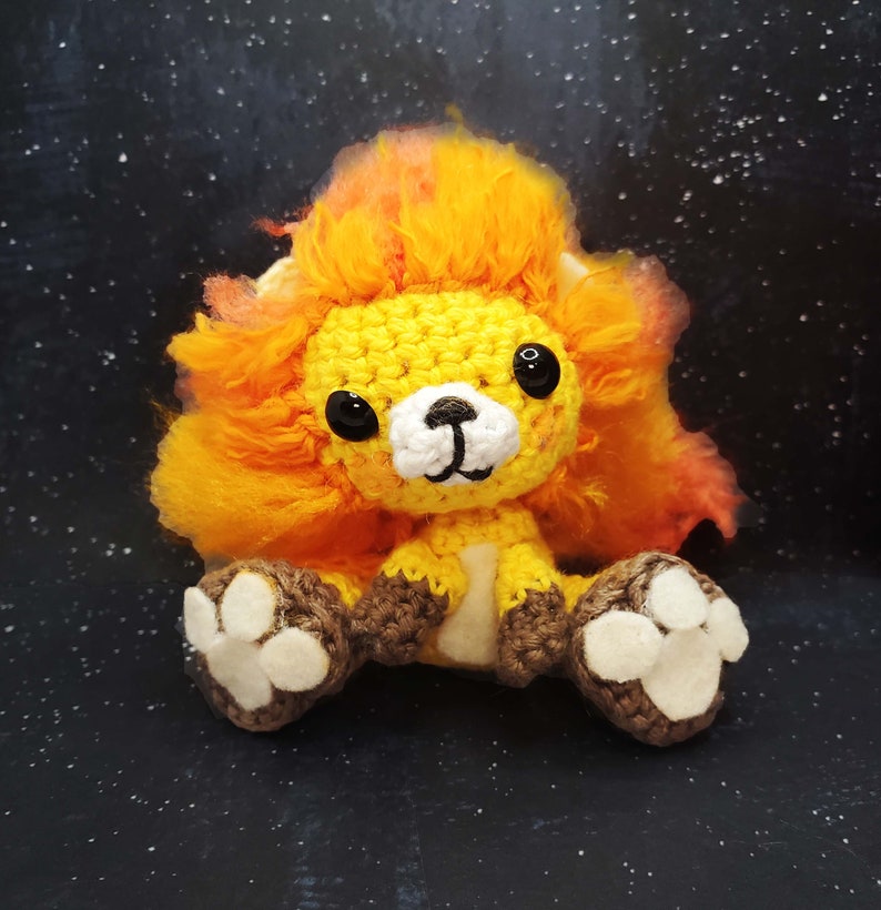 May include: A crocheted lion plush toy with a bright orange mane, yellow body, and white face. The toy has brown crocheted paws with white paw pads.