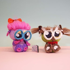 May include: Two handmade crochet owl dolls with large eyes and glasses. One owl has pink hair and a bow, the other has brown hair. A tiny book sits between them. The owls are on a pink surface with a light green background.