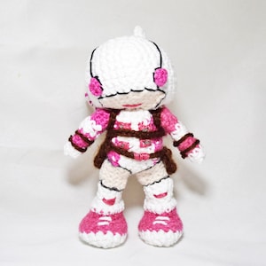 May include: White crocheted doll with pink and brown details. The doll has a white helmet with pink accents and is wearing a pink and brown outfit. The doll is standing on a white surface.