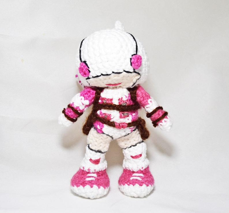 May include: White crocheted doll with pink and brown details. The doll has a white helmet with pink accents and is wearing a pink and brown outfit. The doll is standing on a white surface.