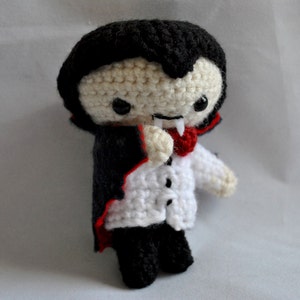 May include: A crocheted plush toy vampire with a black cape, white shirt, and red bow tie. The toy has a black face with white eyes and a red mouth with fangs.
