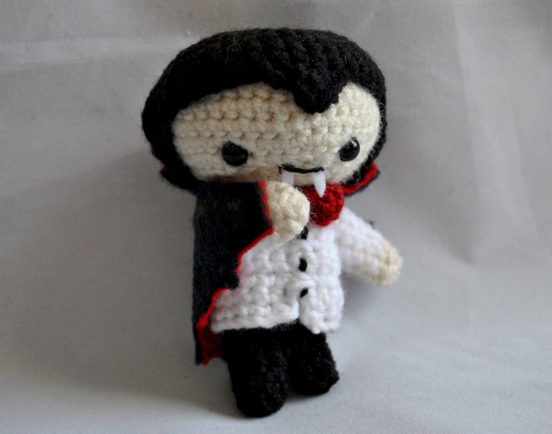 May include: A crocheted plush toy vampire with a black cape, white shirt, and red bow tie. The toy has a black face with white eyes and a red mouth with fangs.
