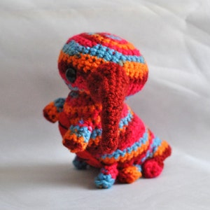 May include: A crocheted stuffed animal in the shape of a creature with a red, orange, and blue striped body and a red, crocheted trunk. The creature has a single black eye.