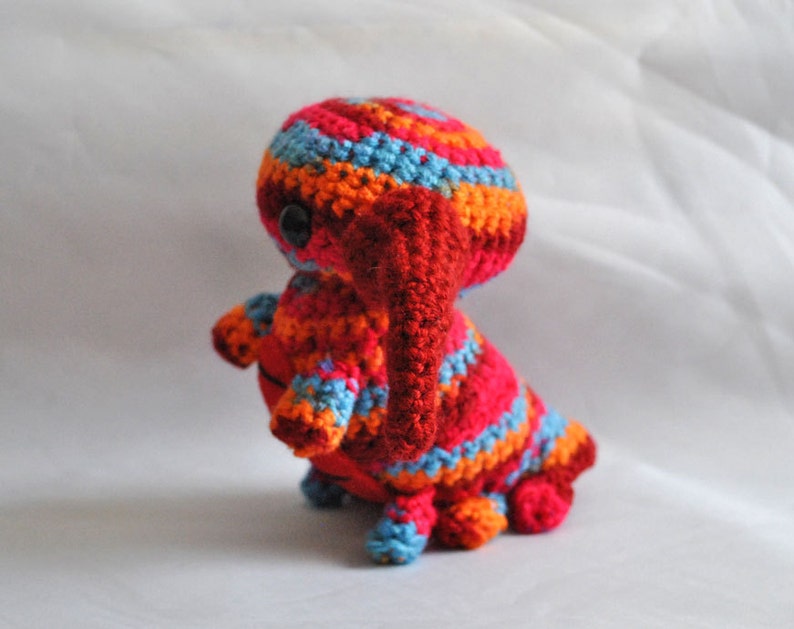 May include: A crocheted stuffed animal in the shape of a creature with a red, orange, and blue striped body and a red, crocheted trunk. The creature has a single black eye.