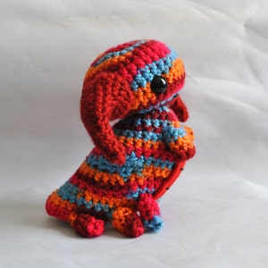 May include: A crocheted stuffed toy in the shape of a creature with a long, pointed snout, large eyes, and a striped body. The toy is made in shades of red, orange, blue, and white.