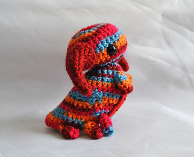 May include: A crocheted stuffed toy in the shape of a creature with a long, pointed snout, large eyes, and a striped body. The toy is made in shades of red, orange, blue, and white.