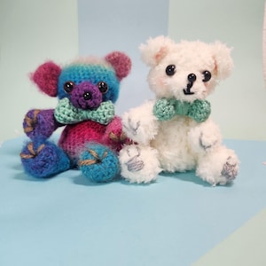 May include: Two handmade crochet teddy bears. One bear is white with a green bow tie, the other is colorful with blue, purple, and pink hues and a green bow tie. The bears are sitting on a light blue surface.