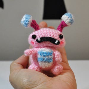 May include: A pink crocheted amigurumi toy with blue and white accents. The toy has a black moustache and large black eyes.
