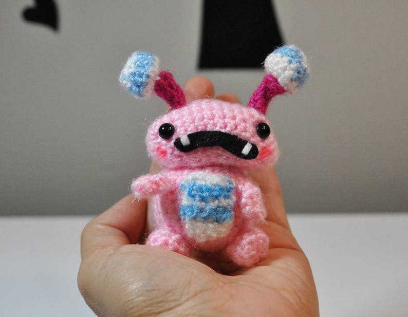 May include: A pink crocheted amigurumi toy with blue and white accents. The toy has a black moustache and large black eyes.