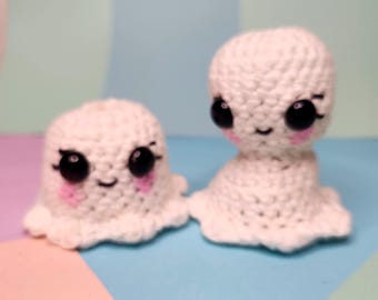 May include: Two handmade white crochet ghost figures with black eyes, pink cheeks, and smiling mouths. The ghosts have a rounded shape with a ruffled bottom edge. They are displayed on a pastel-colored background.
